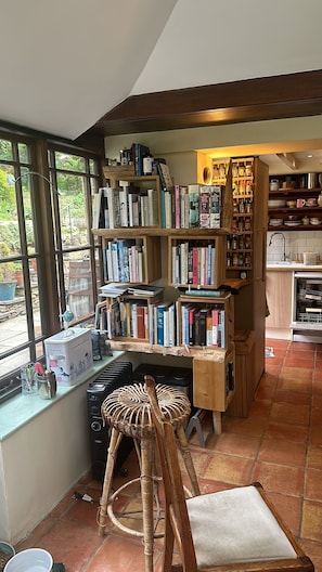 A cozy kitchen with a large window, wooden bookshelves, a wicker stool, and a tiled floor.