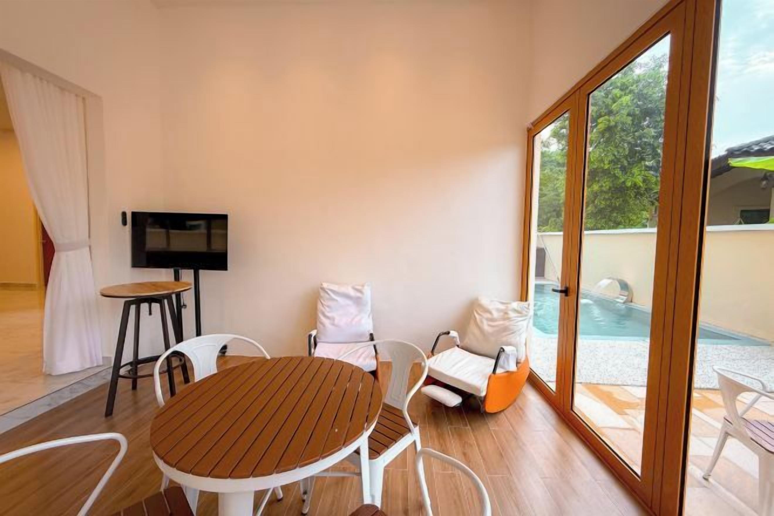 A room with a wooden table, white chairs, a TV, and a view of a swimming pool through large glass doors.