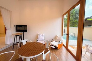 Une pièce avec une table en bois, des chaises blanches, un téléviseur et une vue sur une piscine, visible à travers de grandes portes vitrées.