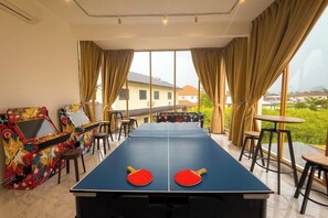 Une table de ping-pong avec deux raquettes, une borne d’arcade aux couleurs vives, et une vue sur une maison à travers de grandes fenêtres.