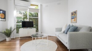 A modern living room with a grey sofa, a glass coffee table, and a flat-screen TV on a white cabinet. - Seaside 1-bed Stay Near Cafes & Attractions (Melbourne)