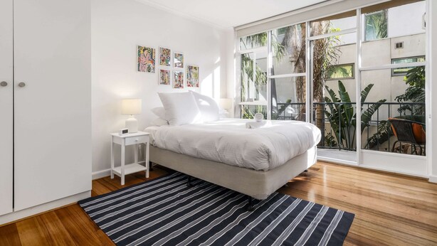 A bedroom with a bed, a nightstand, a lamp, and a view of a balcony with plants. - Seaside 1-bed Stay Near Cafes & Attractions (Melbourne)