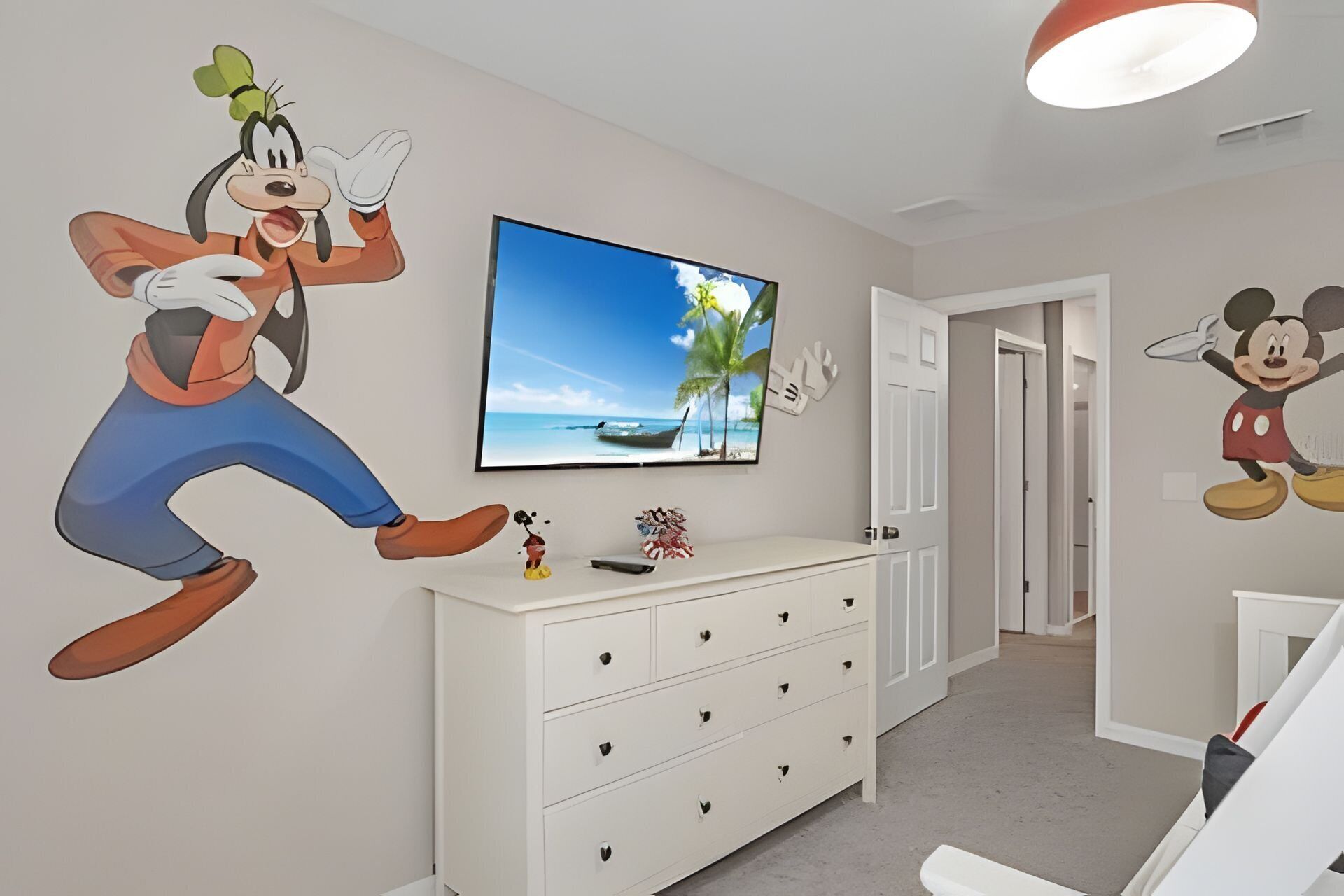 A room with a television mounted on the wall displaying a beach scene, a white dresser with drawers, and cartoon character wall decals.