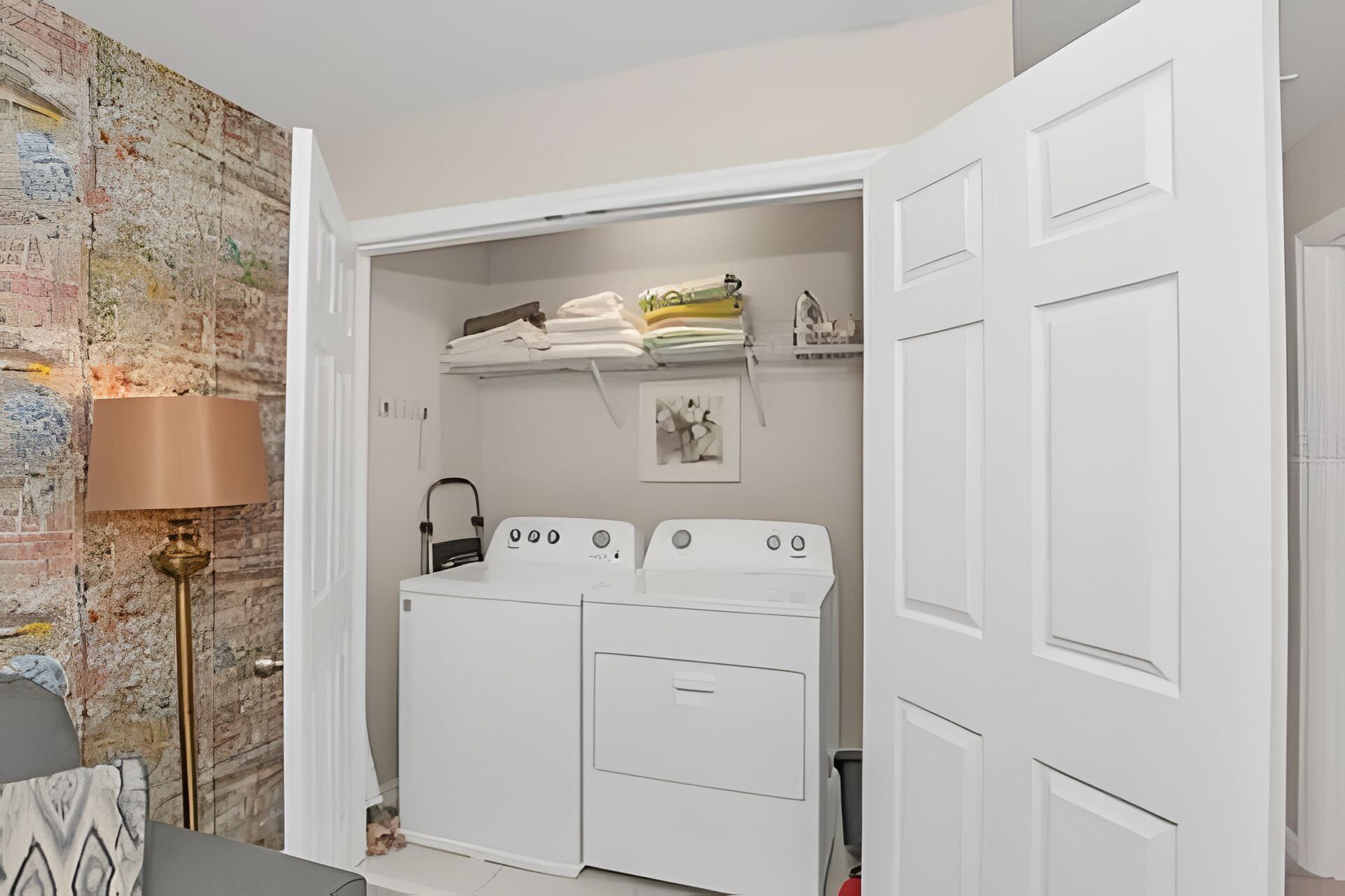 A laundry room with a white washing machine and dryer, a shelf with folded towels, and a lamp.