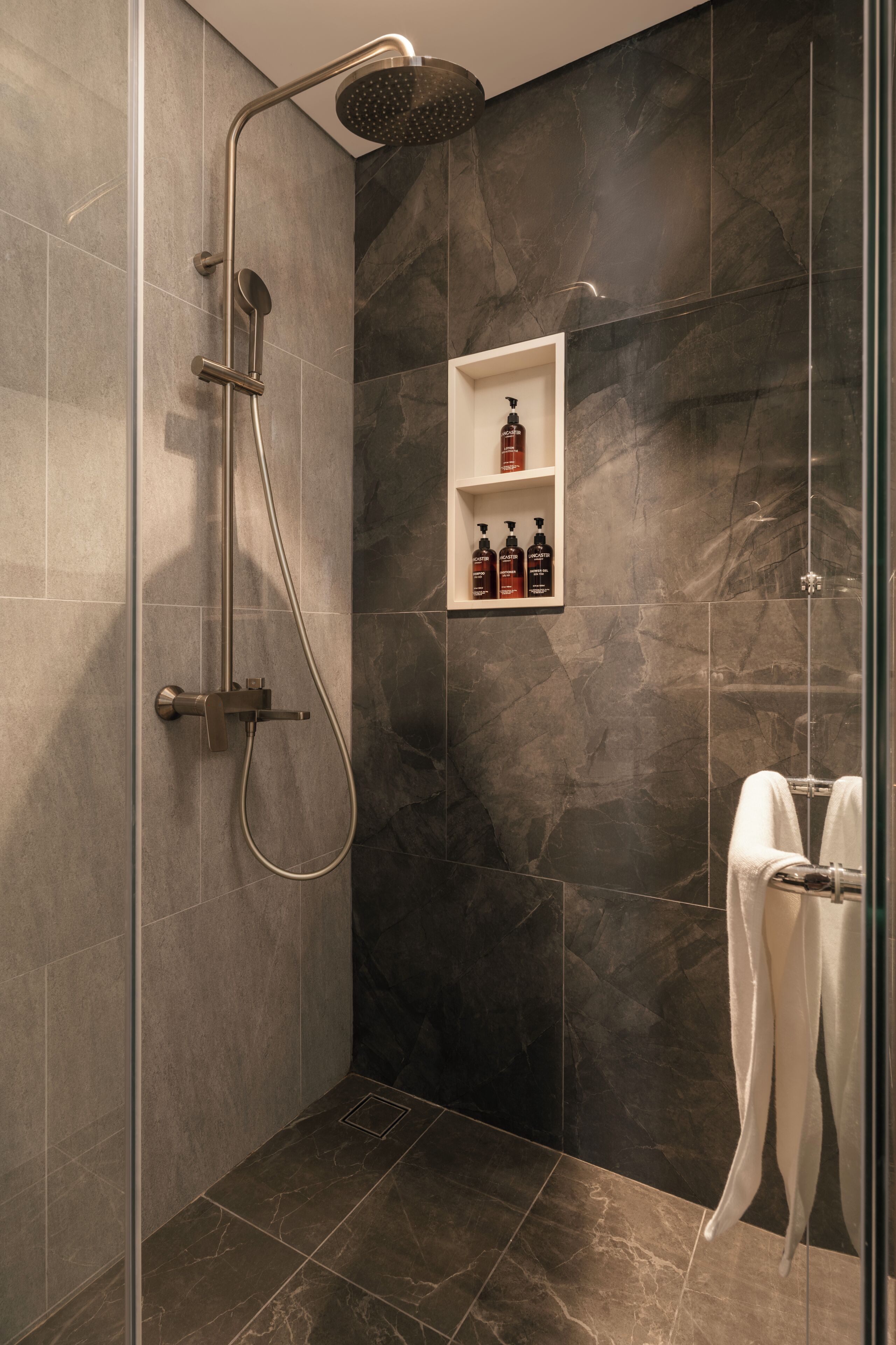 Una ducha moderna con cabezal de ducha manual y llave de pared, zona de ducha con mampara de vidrio y un estante pequeño con artículos de aseo.