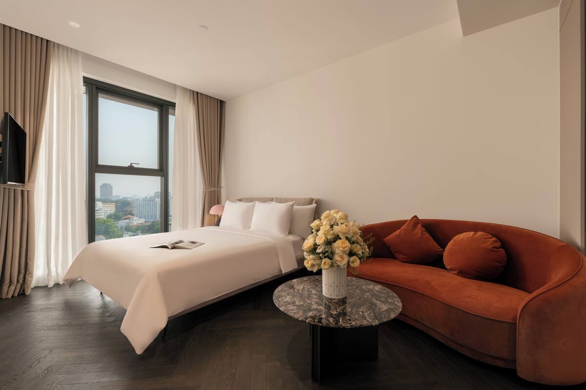 Una habitación de hotel moderna con una cama grande, un sofá, una mesa de centro redonda con un florero y vistas a la ciudad a través de un ventanal.