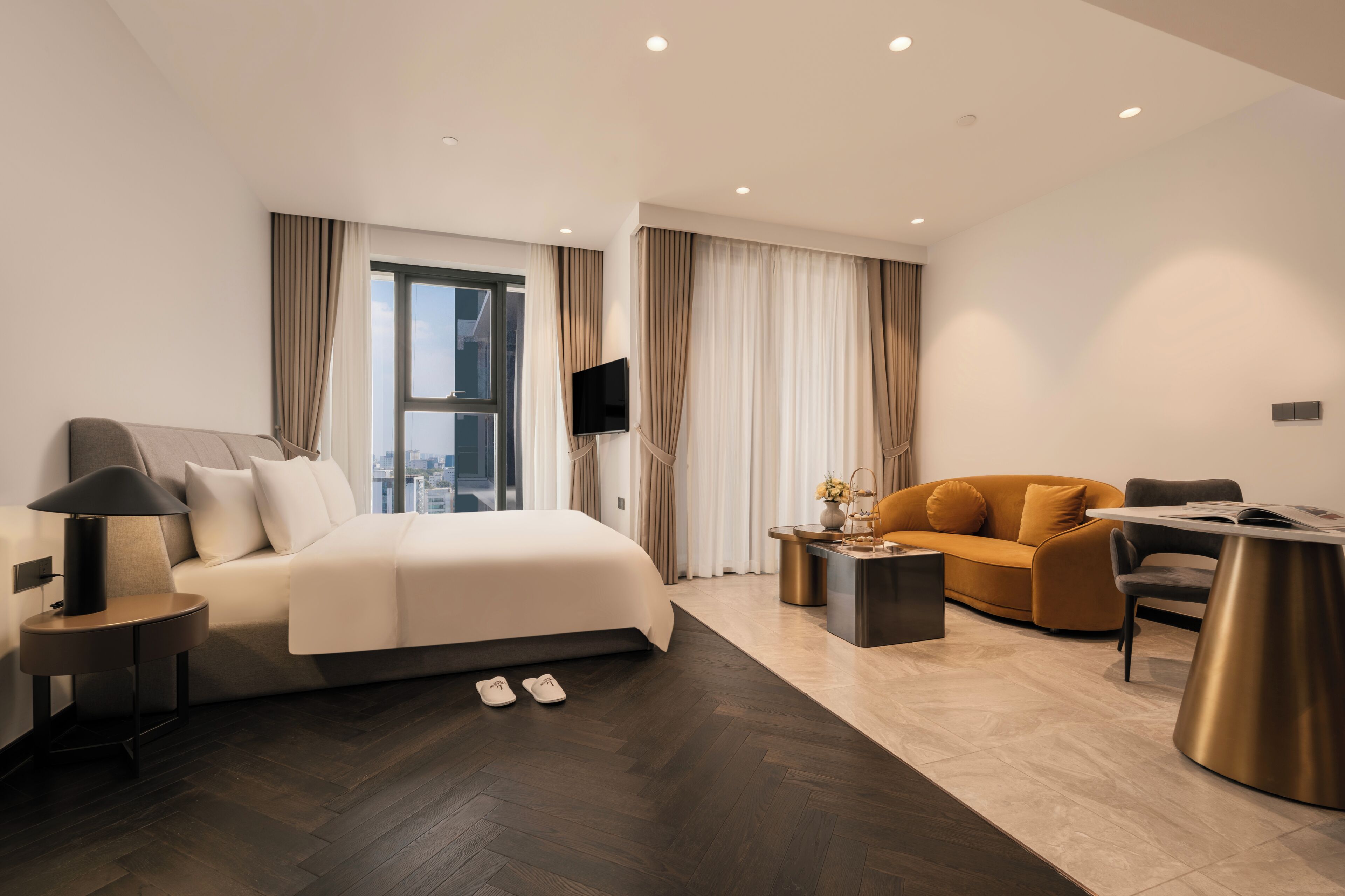 A modern hotel room with a bed, a sofa, a desk, a TV, and a window with curtains.