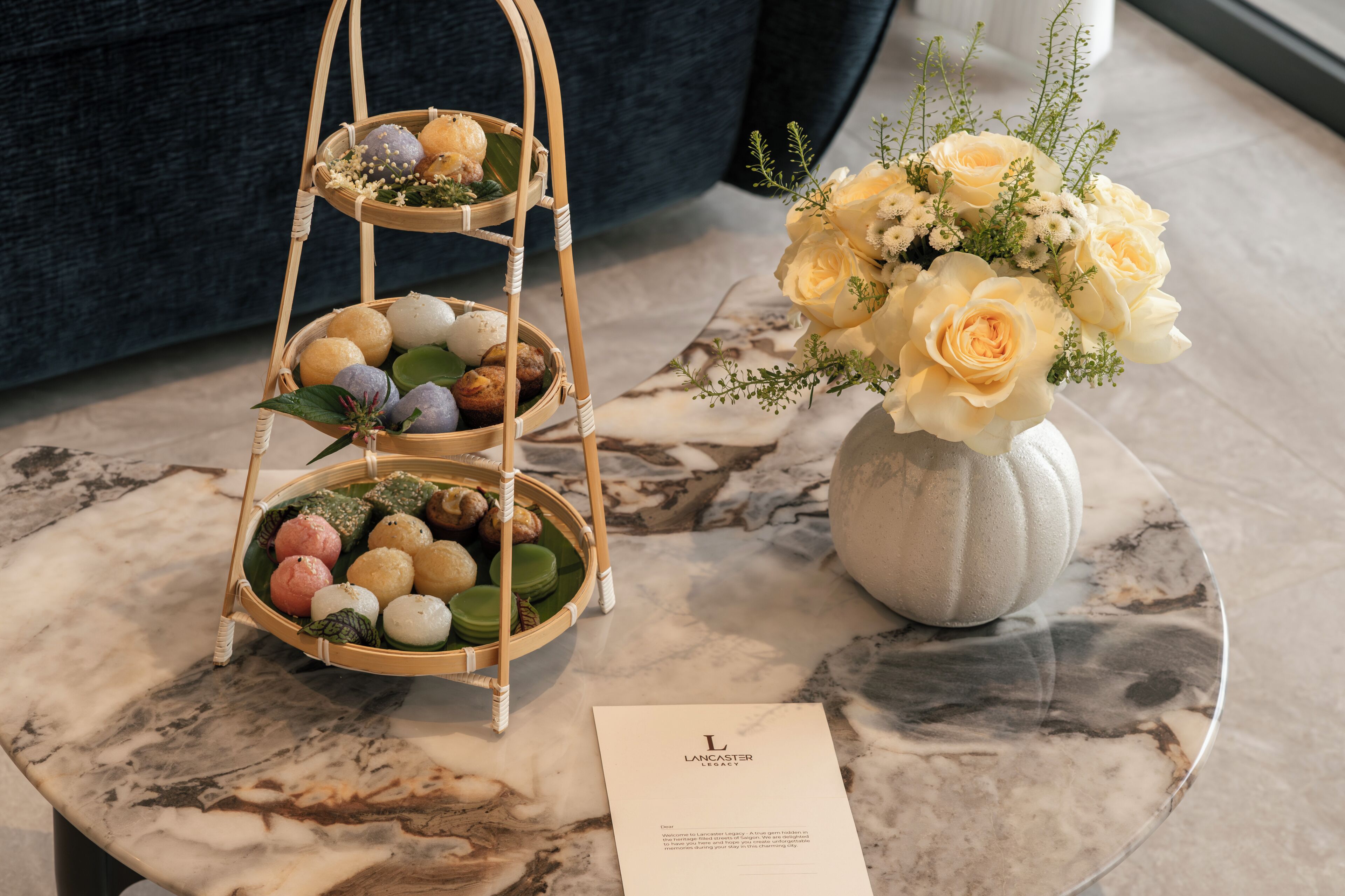 Una mesa de mármol con una cesta escalonada de diversos pasteles, un jarrón de flores y una tarjeta de presentación.