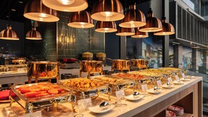 Un bufé con diversos alimentos, un interior moderno con lámparas colgantes de cobre y vistas al exterior a través de amplios ventanales.