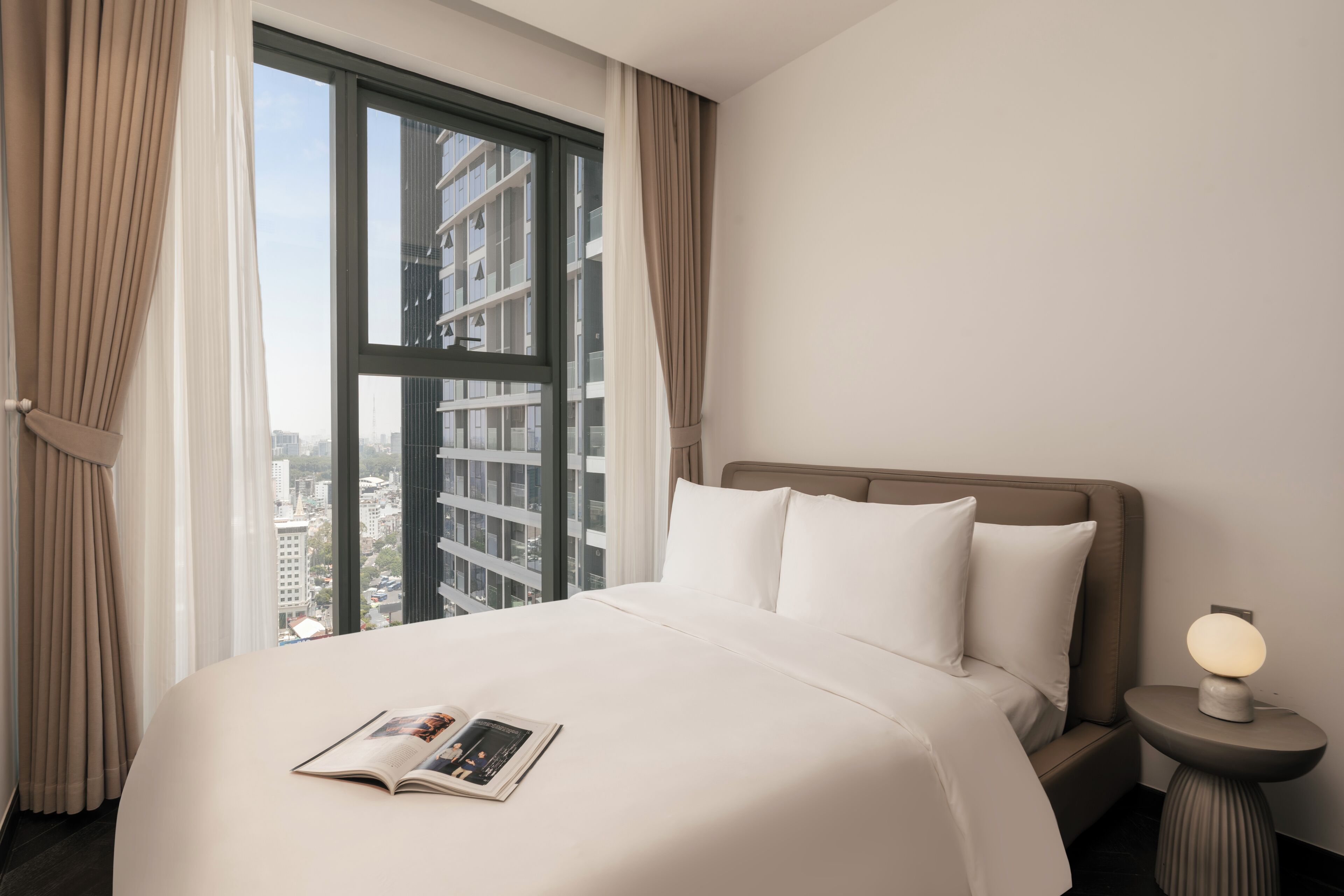 A hotel room with a large bed, a nightstand with a lamp, and a view of a cityscape through a large window.