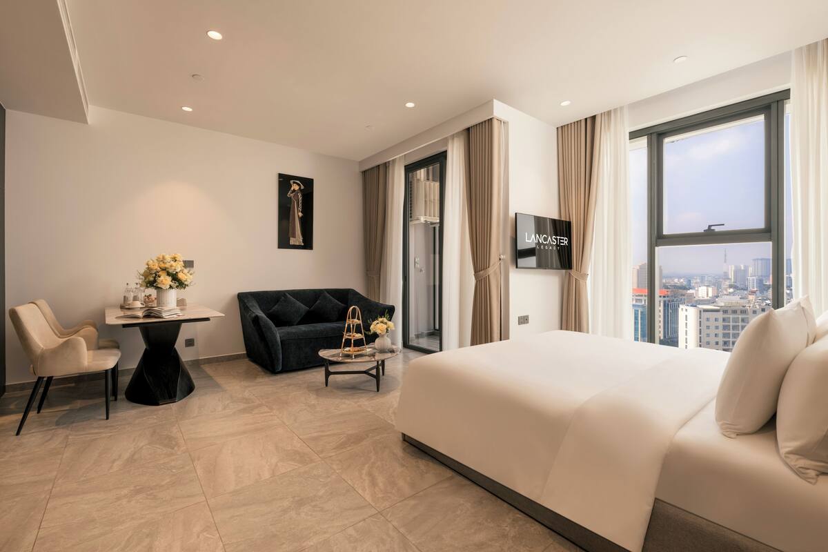 Habitación de hotel con cama, sofá, mesa con un florero y televisión, y vista a la ciudad.