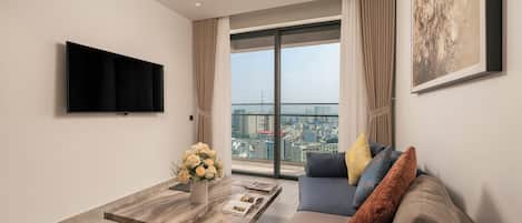 A modern living room with a flat-screen TV, a sofa with cushions, a coffee table with a vase of flowers, and a large window with a city view.