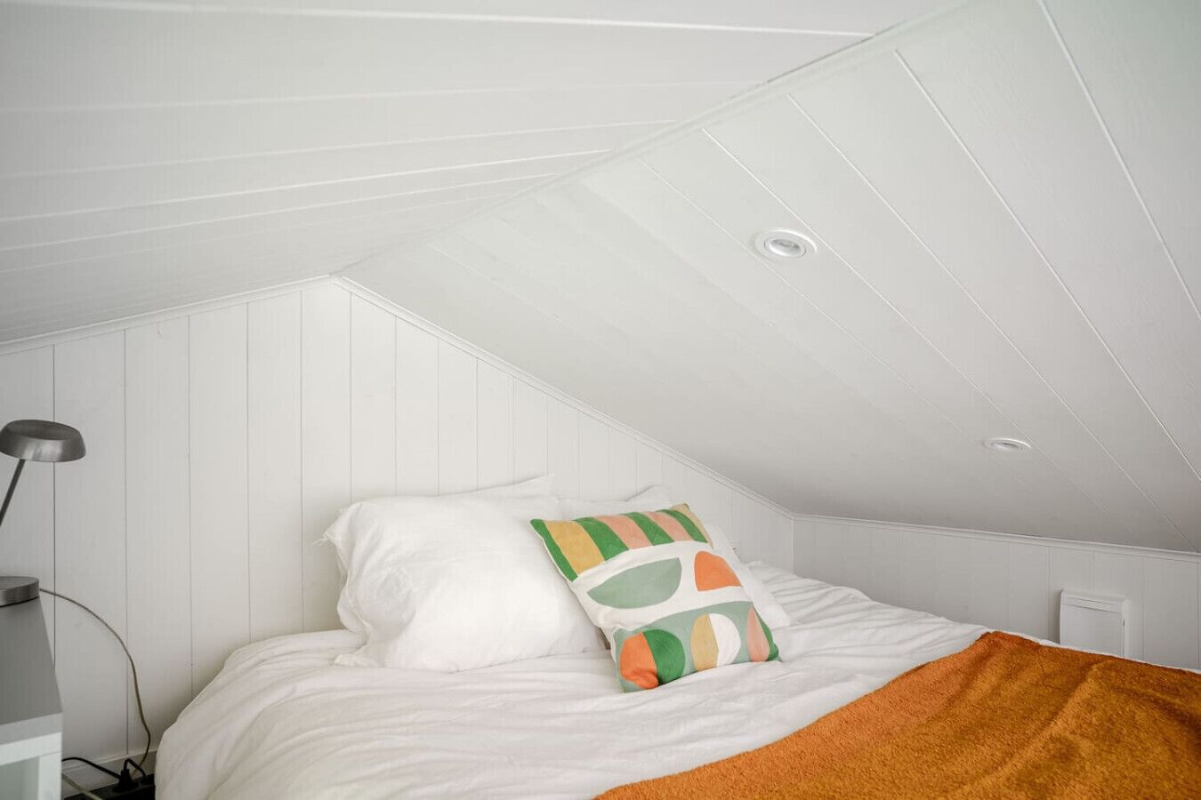 A bedroom with a sloped ceiling, a bed with white bedding, a colorful pillow, and a brown throw blanket.