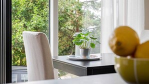 Une fenêtre donnant sur des arbres, une chaise, une plante en pot et un saladier de fruits sur une table.