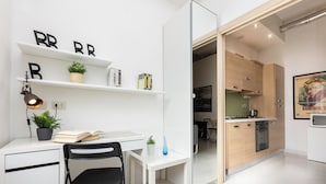 A modern, well-lit room with a white desk, black chair, and a small table with a plant and a vase. Shelves above the desk hold books and decorative items. A kitchen area is visible through an open door.