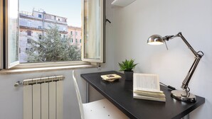 A room with a black desk, a window with a view of buildings, a radiator, a white chair, a desk lamp, a plant, and some books.