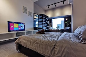 Ein modernes Schlafzimmer mit Bett, Fernseher, Schreibtisch und einem Fenster mit Blick auf die Stadt.