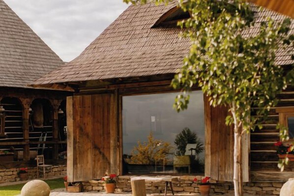 A rustic log cabin with a large glass window, stone walls, and a wooden deck. - ECHOES OF VLĂDICU (Cluj-Napoca)