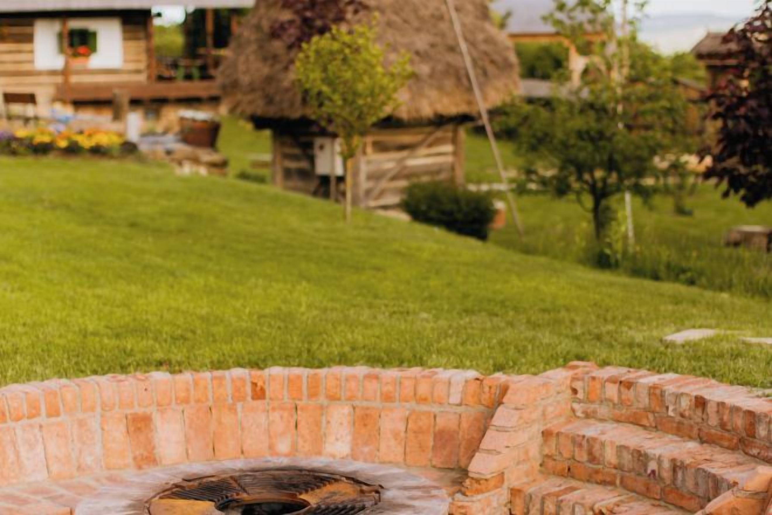 A brick outdoor fireplace surrounded by a grassy area with a thatched-roof structure in the background.