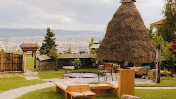 A thatched-roof structure, picnic tables, and a stone wall in a garden setting with a city view.