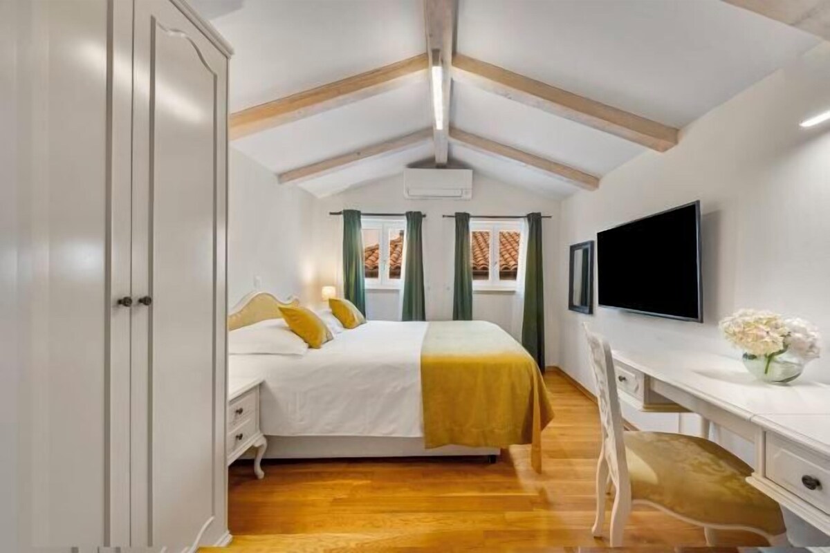 Een slaapkamer met een bed, een bureau, een stoel, een televisie en een raam met gordijnen.