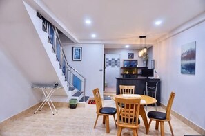 Un espace repas avec une table ronde et des chaises, un escalier et, au fond, une cuisine.