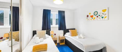 A hotel room with two beds, a mirror, a window with curtains, a bedside table with a lamp, and a wall art piece.