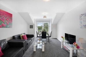 A modern living room with a glass coffee table, black leather sofas, a flat-screen TV, and a dining area with chairs and a table.