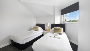 A hotel room with two single beds, a nightstand with a lamp, a window with a view, and a small table with a fruit bowl.