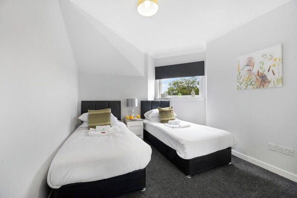 A hotel room with two beds, a window with a view, a painting on the wall, and a small table with a lamp.
