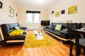 A living room with two black sofas, a glass coffee table, a yellow rug, and a television.