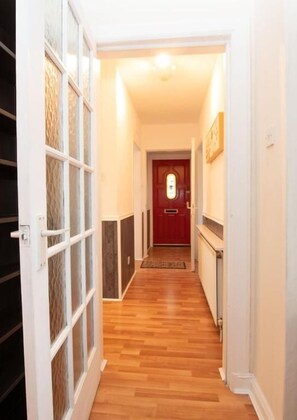A hallway with a wooden floor, a red door at the end, and a white wall with a framed picture.