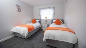 Une chambre avec deux lits simples, chacun agrémenté de draps blancs et de touches de couleur orange, une petite table de chevet avec un vase, et une fenêtre ornée de rideaux à motifs.