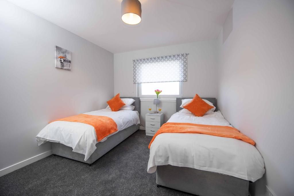 A bedroom with two beds, a window with a patterned blind, a small bedside table with a vase, and a wall-mounted artwork.