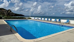 Ein Swimmingpool mit blauen Fliesen, umgeben von Liegestühlen, mit Blick auf das Meer und einen bewölkten Himmel.