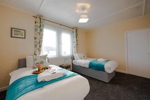 A hotel room with two beds, a window with curtains, a bedside table with a lamp, and a door.