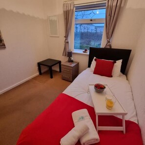 Ein Hotelzimmer mit Bett, Nachttisch mit Lampe, Fenster mit Vorhängen, kleinem Tisch mit Frühstücksbrett und einem Handtuch auf dem Boden.