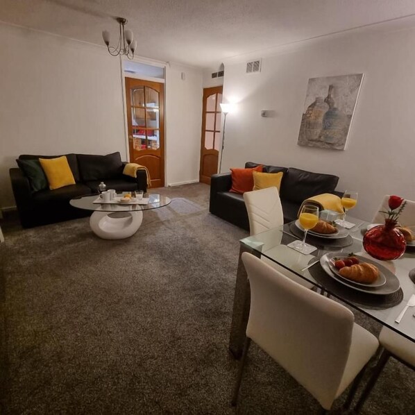 A living room with a glass coffee table, two sofas with cushions, a dining table with breakfast items, and a door leading to another room.