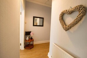 Un couloir doté d’un plancher en bois, une petite table avec une lampe et des fleurs, un miroir encadré accroché au mur et un décor en forme de cœur.