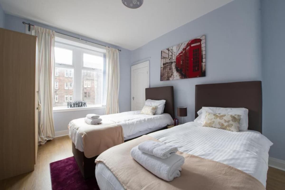 A hotel room with two beds, a window with curtains, a red telephone booth painting, and a view of buildings outside.