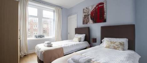 A hotel room with two beds, a window with curtains, a red telephone booth painting, and a view of buildings outside.