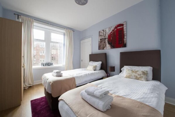 Une chambre d’hôtel avec deux lits, une fenêtre avec des rideaux, un tableau représentant une cabine téléphonique rouge, et une vue sur les bâtiments à l’extérieur.