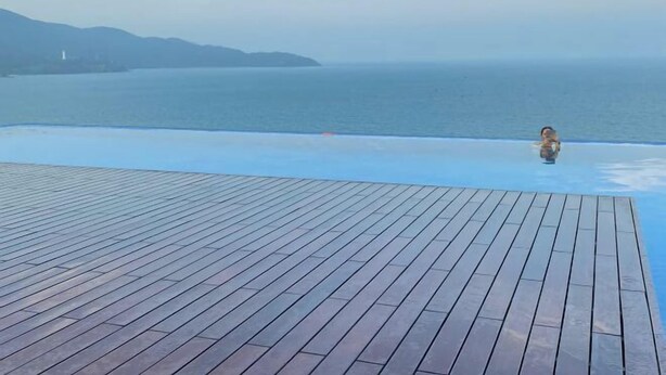 Een overloopzwembad met een houten terras en uitzicht op de zee en verre heuvels.
