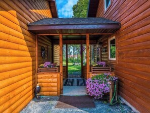 Una cabaña de madera con un porche cubierto, una puerta que da a un jardín y jardineras con flores moradas.