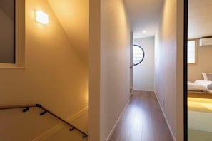 A narrow hallway with a wooden floor, a handrail on the left, and a bed with white bedding in the right room.