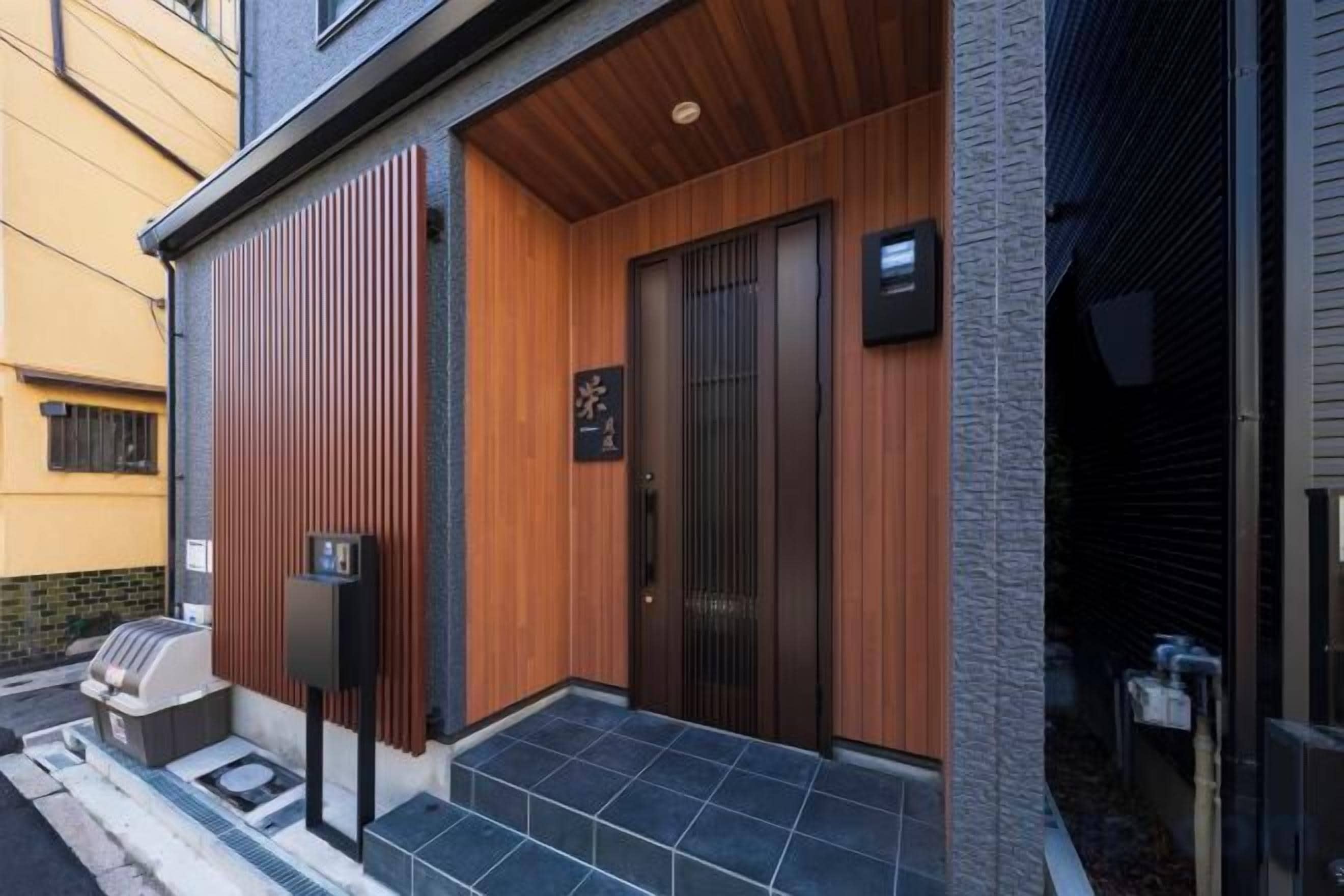 A building entrance with a wooden door, a black mailbox, and a small outdoor seating area.