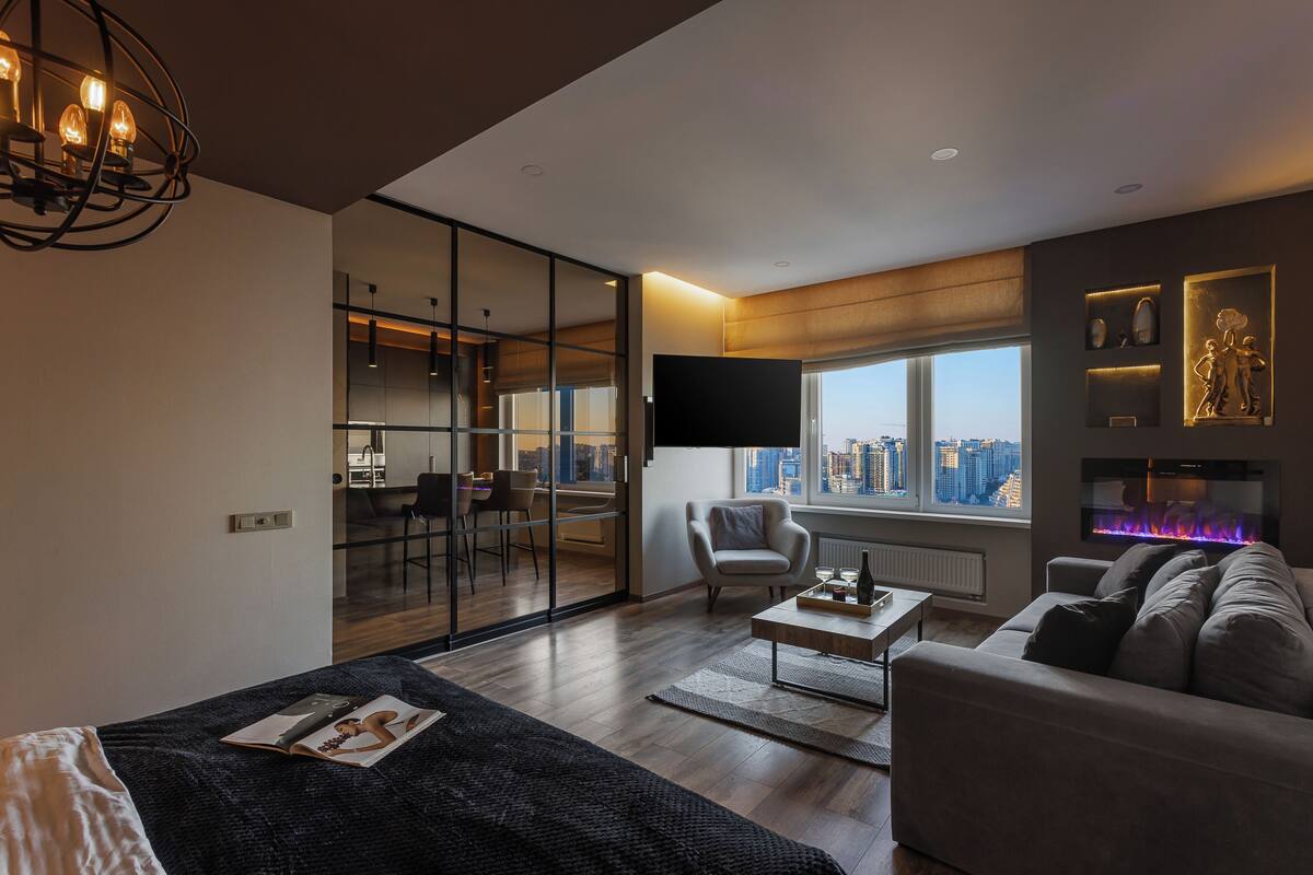 A modern living room with a sofa, coffee table, and a fireplace. There is a TV mounted on the wall and a large window offering a city view.