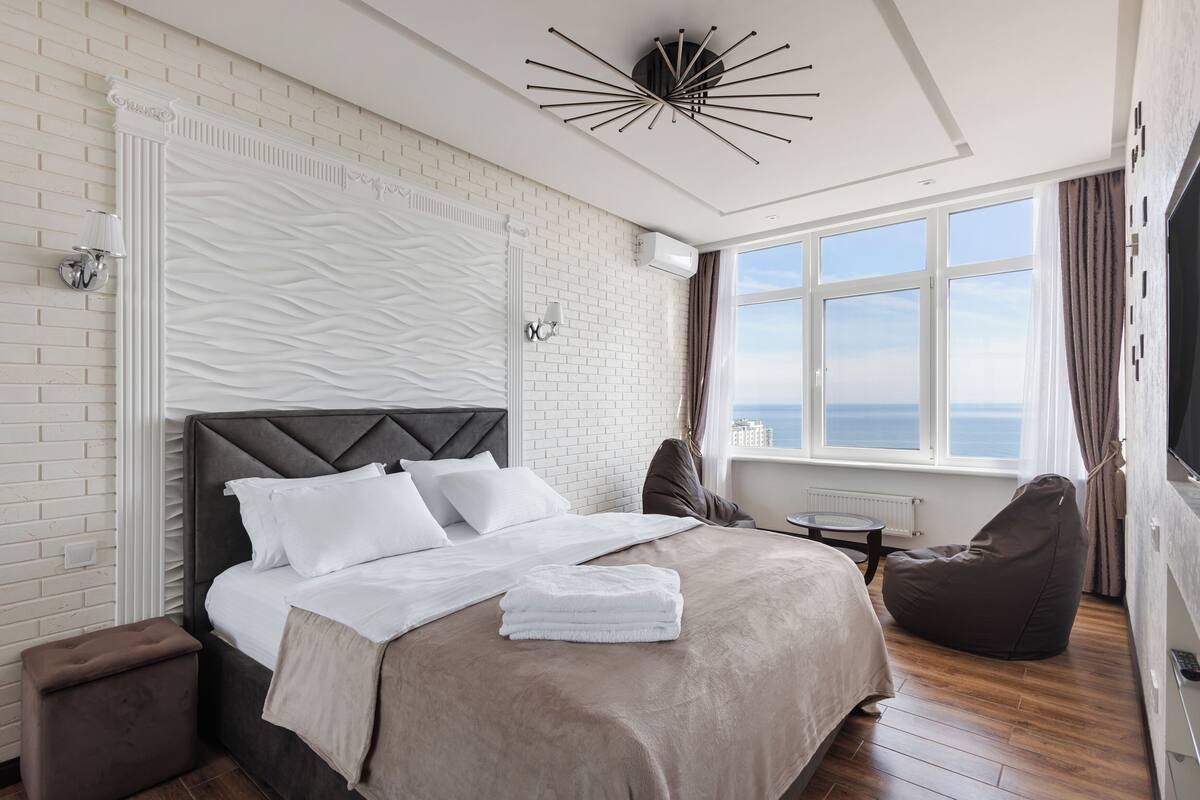 A modern hotel room with a large bed, a view of the sea, a unique ceiling light, and a cozy seating area.