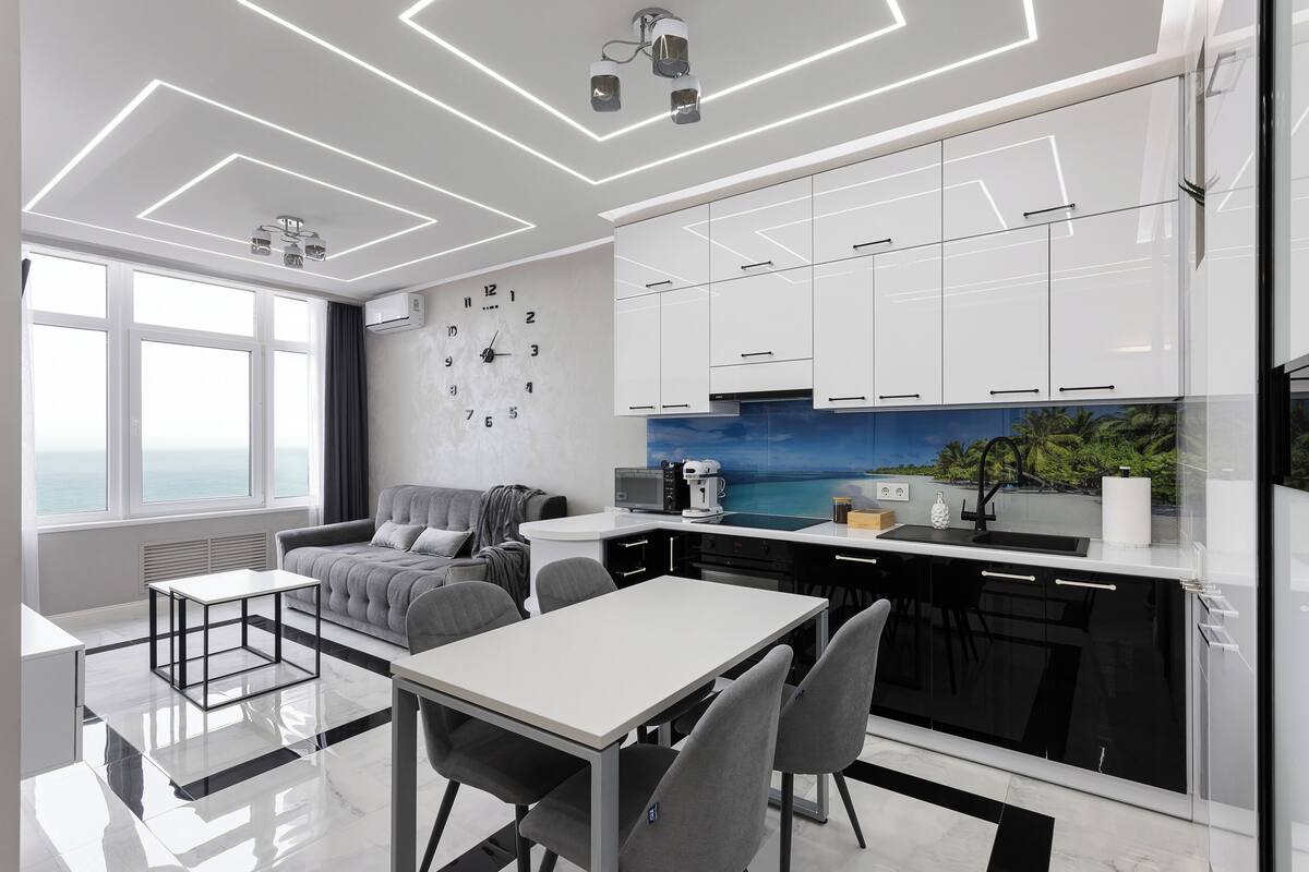 A modern kitchen with white cabinets, a dining area with a table and chairs, a sofa, and a large window with a view.