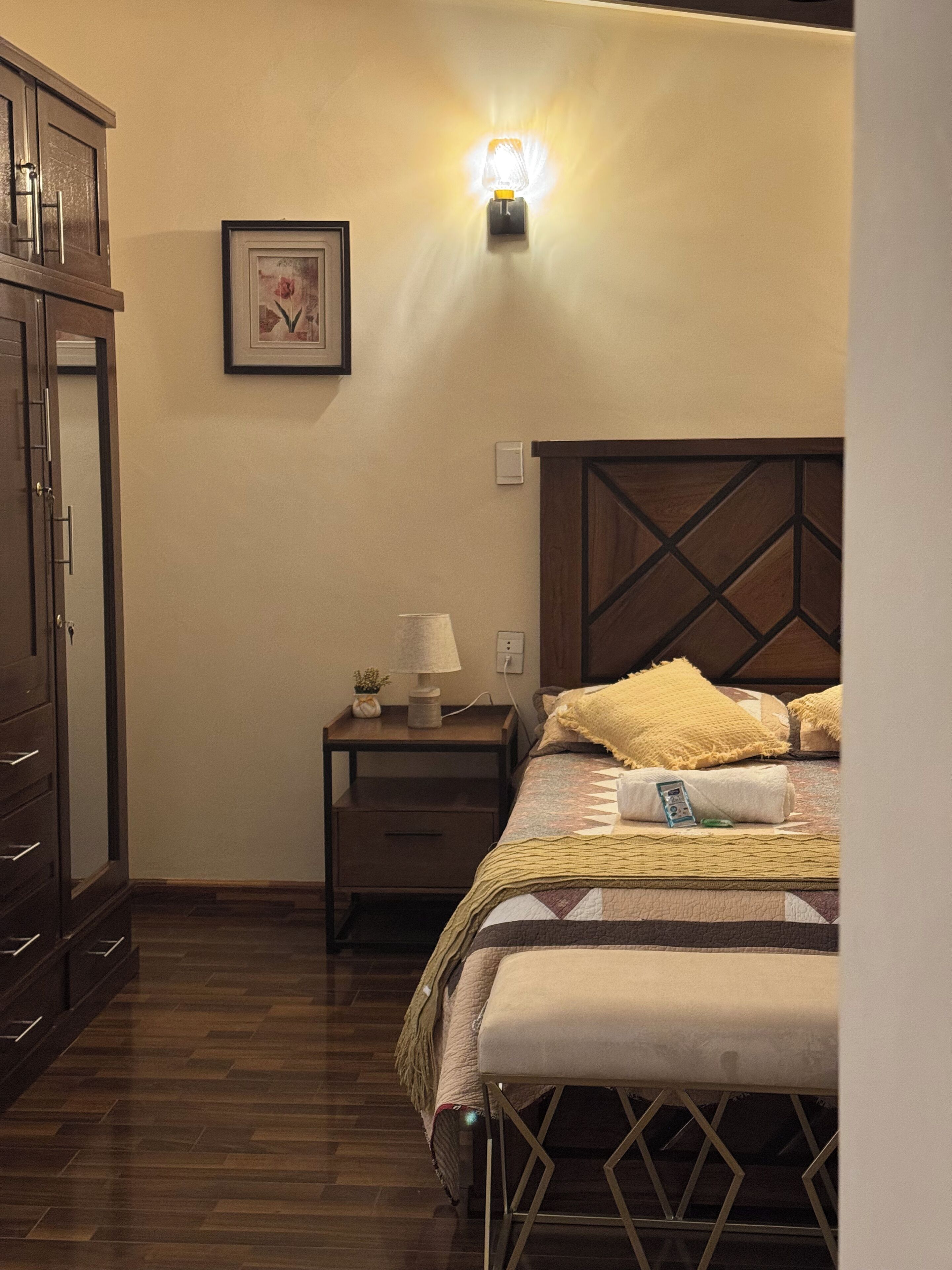 A bedroom with a wooden headboard, a bedside table with a lamp, a framed picture on the wall, and a wooden wardrobe.